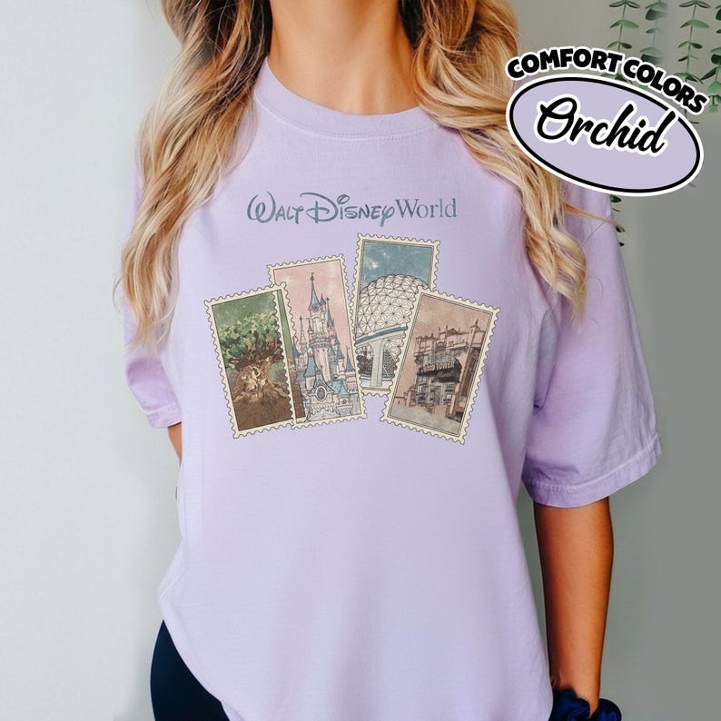 Op de afbeelding: Een lichtpaars T-shirt met de tekst "Walt Disney World" op de voorkant. Het shirt heeft vier postzegelachtige afbeeldingen van Disney-attracties. De woorden "Comfort Colors" en "Orchid" staan rechtsboven.