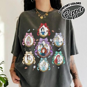 May include: Dark gray t-shirt featuring framed illustrations of animated princesses. Each frame is adorned with a bow and contains a princess. The shirt has the text "Comfort Colors Pepper" in a circular design.
