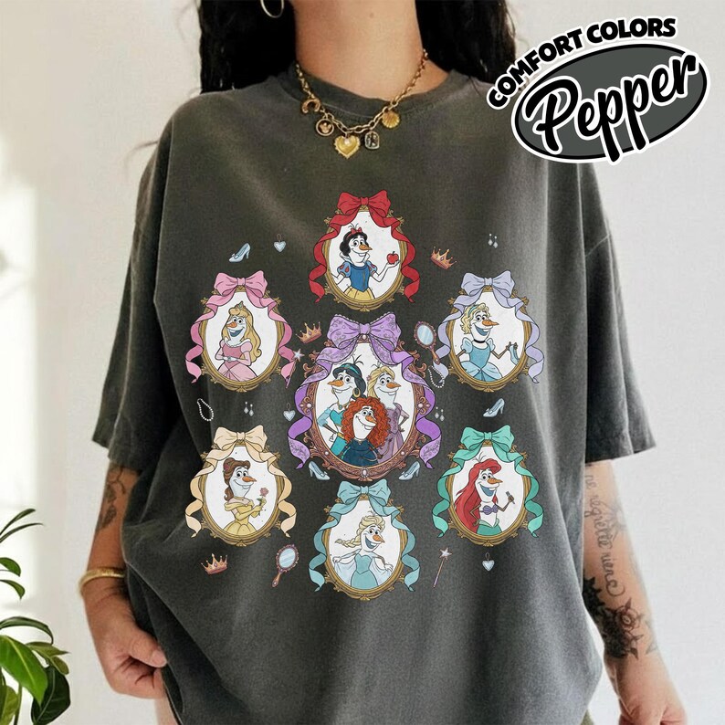 May include: Dark gray t-shirt featuring framed illustrations of animated princesses. Each frame is adorned with a bow and contains a princess. The shirt has the text "Comfort Colors Pepper" in a circular design.