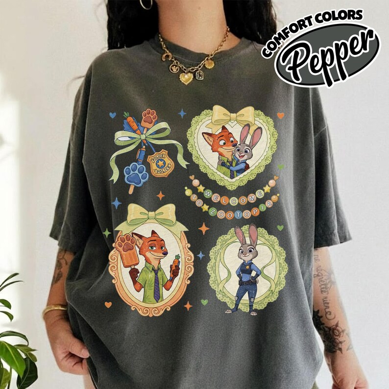 May include: A charcoal gray t-shirt featuring a graphic print of Zootopia characters Nick Wilde and Judy Hopps. The design includes ornate frames, bows, a police badge, a paw print, and the text Welcome to Zootopia in a circular arrangement.
