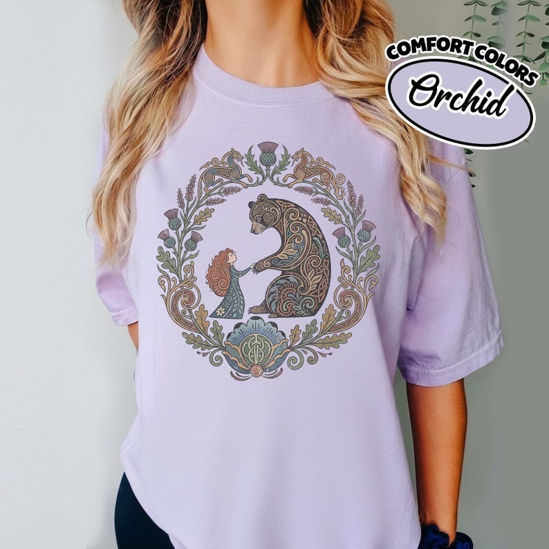 May include: A light purple t-shirt with a circular graphic featuring a bear and a girl, surrounded by floral elements. The graphic is in shades of brown, green, and blue. The text "COMFORT COLORS Orchid" is in a white oval.