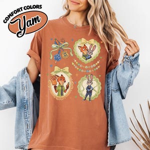 May include: A rust-colored short-sleeve t-shirt featuring a graphic print of Zootopia characters Nick Wilde and Judy Hopps. The design includes four framed illustrations with bows, a police badge, a popsicle, and the text Welcome to Zootopia.