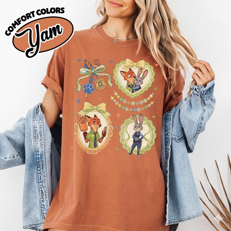 May include: A rust-colored short-sleeve t-shirt featuring a graphic print of Zootopia characters Nick Wilde and Judy Hopps. The design includes four framed illustrations with bows, a police badge, a popsicle, and the text Welcome to Zootopia.