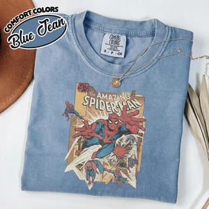 K&ouml;nnte beinhalten: Blaues Comfort Colors T-Shirt mit einem Vintage-Comic-Grafik von Spider-Man. Das Shirt hat einen Rundhalsausschnitt und kurze &Auml;rmel. Eine silberne Halskette mit Anh&auml;nger ist &uuml;ber dem Shirt drapiert.