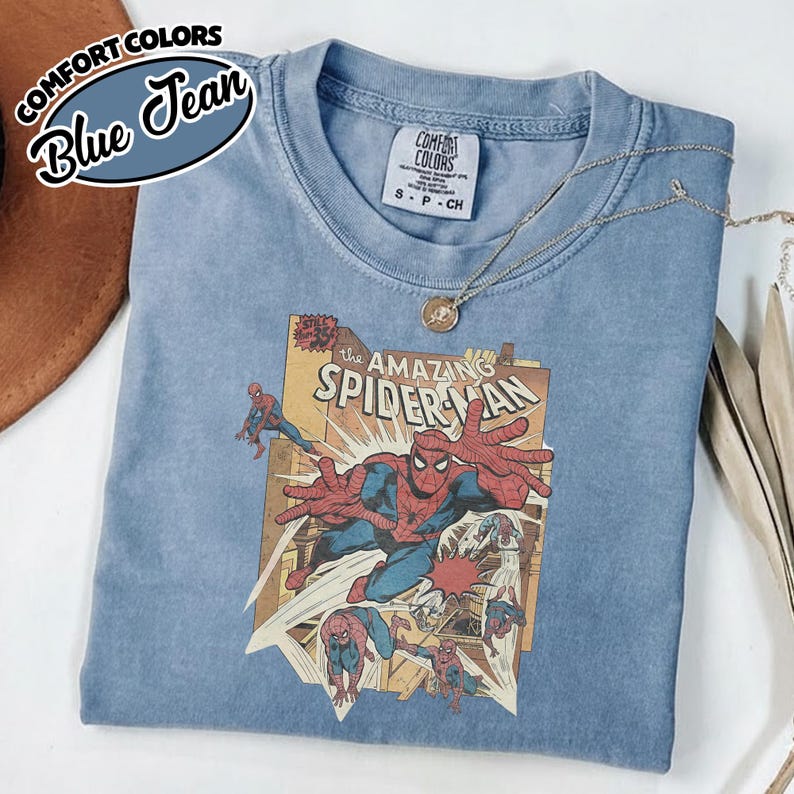 K&ouml;nnte beinhalten: Blaues Comfort Colors T-Shirt mit einem Vintage-Comic-Grafik von Spider-Man. Das Shirt hat einen Rundhalsausschnitt und kurze &Auml;rmel. Eine silberne Halskette mit Anh&auml;nger ist &uuml;ber dem Shirt drapiert.