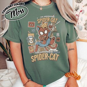 May include: A moss green t-shirt with a graphic of a Spider-Man-inspired cat character. The design includes the text "THE AMEOWZING SPIDER-CAT" and various cartoon cat illustrations. The shirt is a comfort colors style.