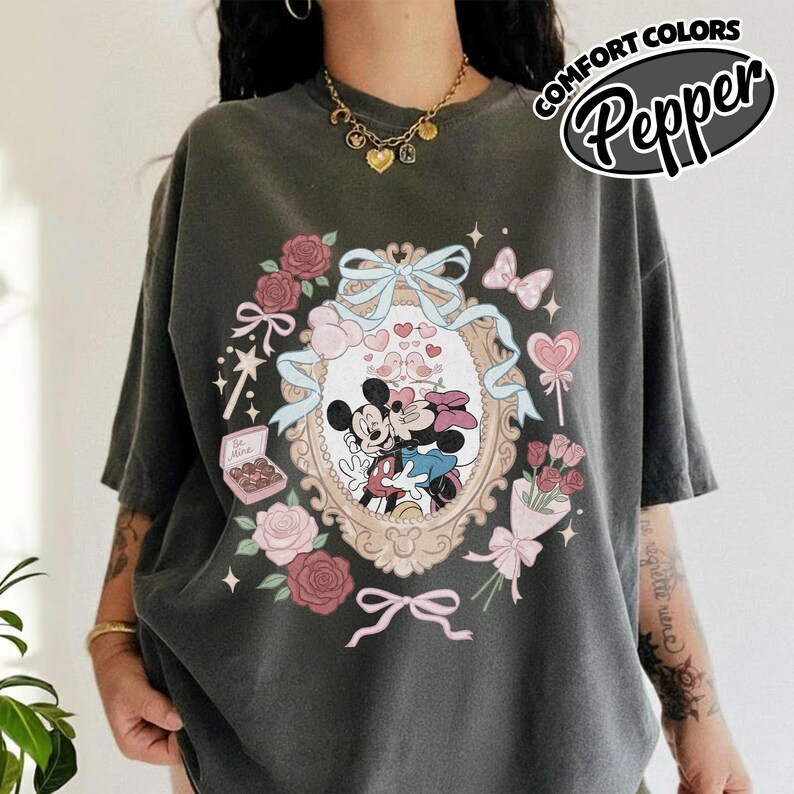 Puede incluir: Una camiseta gris carb&oacute;n con un gr&aacute;fico de Mickey y Minnie Mouse dentro de un marco ovalado ornamentado. El dise&ntilde;o incluye cintas azul claro, rosas rosadas, una caja de bombones, una piruleta y peque&ntilde;as estrellas.