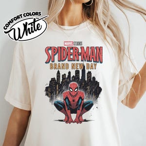 Puede incluir: Una camiseta blanca con un estampado gr&aacute;fico de Spider-Man en cuclillas. Sobre el personaje, el texto dice Marvel Studios Spider-Man Brand New Day en rojo y amarillo. Detr&aacute;s se ve la silueta negra de un horizonte urbano.