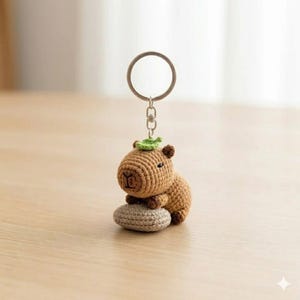 May include: A crochet capybara keychain featuring a brown yarn body, a small green leaf accessory on its head, and a grey crocheted oval base. The figure is attached to a silver-toned metal split ring by a short chain and a lobster clasp.