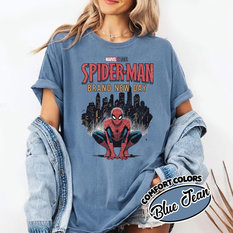 Puede incluir: Una camiseta de color azul mezclilla con un gr&aacute;fico de Spider-Man agachado frente al horizonte de una ciudad. El texto dice Marvel Studios Spider-Man Brand New Day. La prenda tiene mangas cortas y cuello redondo, usada debajo de una chaqueta vaquera.