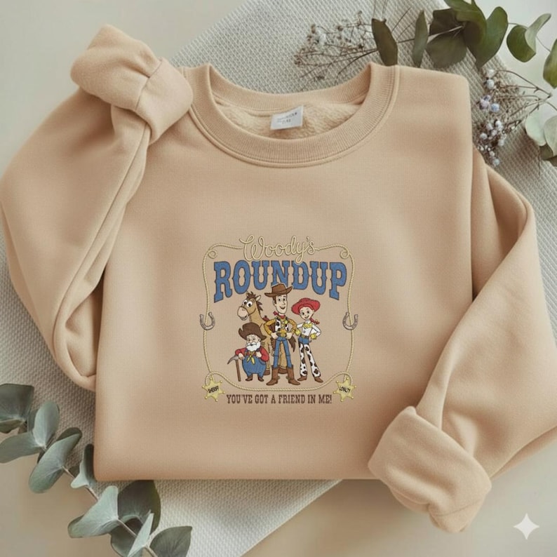 Puede incluir: Una sudadera beige de cuello redondo con un estampado gr&aacute;fico central de Woody, Jessie, Bullseye y Stinky Pete. El texto dice Woody's ROUNDUP y YOU'VE GOT A FRIEND IN ME, enmarcado por un dise&ntilde;o de lazo con dos peque&ntilde;as estrellas de sheriff.
