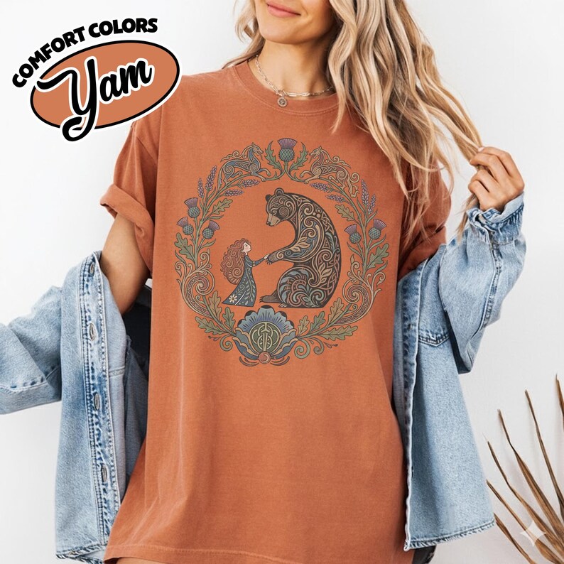 May include: Burnt orange t-shirt with a circular graphic featuring a bear and a girl holding hands, surrounded by a floral wreath. The shirt has the text "COMFORT COLORS" and "Yam" in a brown oval. The shirt has rolled-up sleeves.