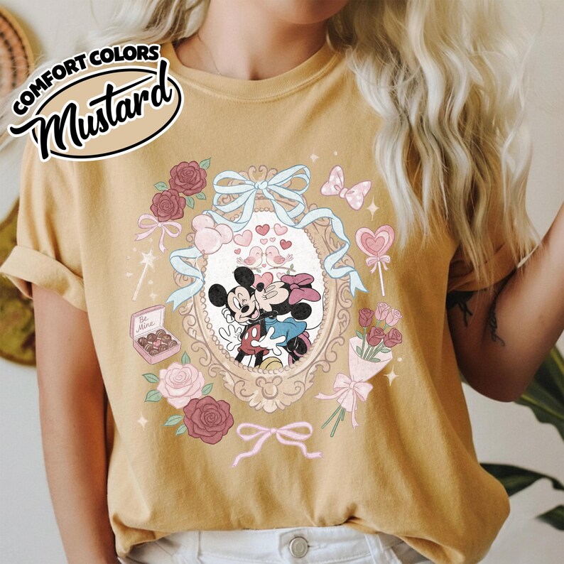 Puede incluir: Una camiseta color amarillo mostaza con un gr&aacute;fico de Mickey y Minnie Mouse bes&aacute;ndose dentro de un marco ovalado ornamentado. El dise&ntilde;o incluye cintas azul claro, rosas rosas y rojas, una caja de bombones con la etiqueta Be Mine y corazones.