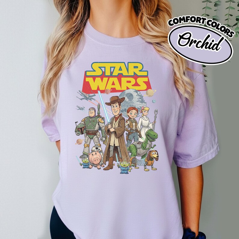 May include: A light purple t-shirt featuring a graphic of Star Wars characters, including Woody, Buzz Lightyear, and others. The Star Wars logo is in yellow and red. The shirt also has the words "Comfort Colors" and "Orchid" printed on it.