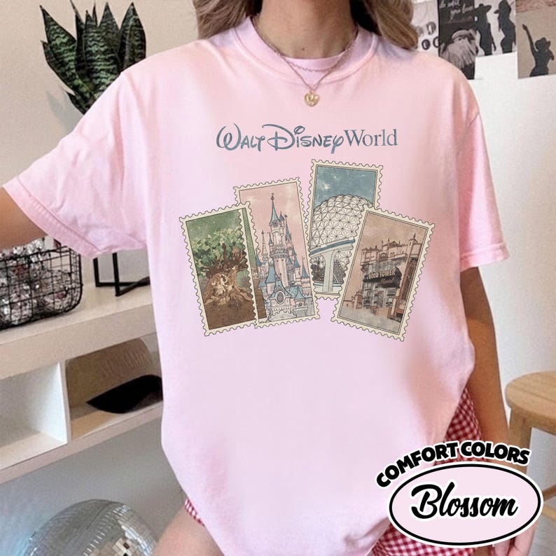Op de afbeelding: Lichtroze T-shirt met de tekst "Walt Disney World" en vier postzegelafbeeldingen van Disney World-bezienswaardigheden. Het shirt heeft ook de tekst "Comfort Colors Blossom" in een ovaal ontwerp.