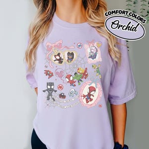 K&ouml;nnte beinhalten: Ein helllila T-Shirt mit einer farbenfrohen Grafik verschiedener Spider-Man-Charaktere und verwandter Elemente. Das Design umfasst gerahmte Portr&auml;ts, Herzen und Spinnennetze. Der Text "COMFORT COLORS" und "Orchid" ist oben rechts aufgedruckt.