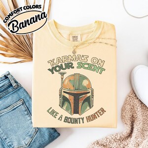 May include: Banana-colored t-shirt with the text "Karma's on Your Scent Like a Bounty Hunter" and a graphic of a helmet. The shirt is part of the Comfort Colors collection. The shirt is folded next to a pair of blue jeans.
