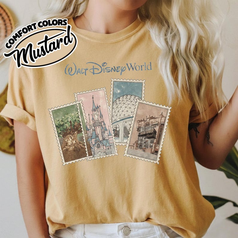 Op de afbeelding: Mosterdkleurig T-shirt met een afbeelding van vier postzegelachtige afbeeldingen van Walt Disney World-attracties. De tekst "Walt Disney World" is lichtblauw gedrukt. Het shirt heeft ook de tekst "Comfort Colors" en "Mustard" in een cirkelvormig ontwerp.