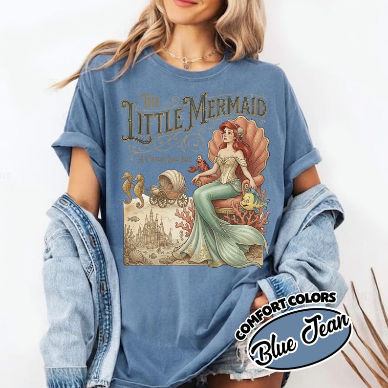 以下が含まれることがあります： ブルーのジーンズComfort Colors Tシャツ。ヴィンテージスタイルのリトルマーメイドのグラフィックが特徴です。デザインには、アリエルが貝殻の玉座に座り、フランダー、セバスチャン、お城が描かれています。シャツは袖をまくっています。