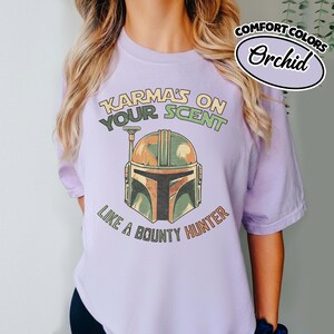 May include: A light purple t-shirt with the text "Karma's on your scent like a bounty hunter" and a graphic of a helmet. The shirt is labeled "Comfort Colors" and "Orchid".