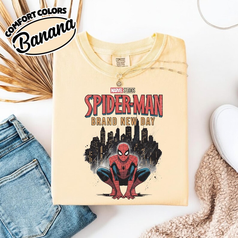 Puede incluir: Una camiseta de color amarillo p&aacute;lido con un gr&aacute;fico de Spider-Man agachado frente a la silueta negra de una ciudad. El texto dice Marvel Studios Spider-Man Brand New Day. La camiseta tiene una etiqueta de la marca Comfort Colors en el cuello.