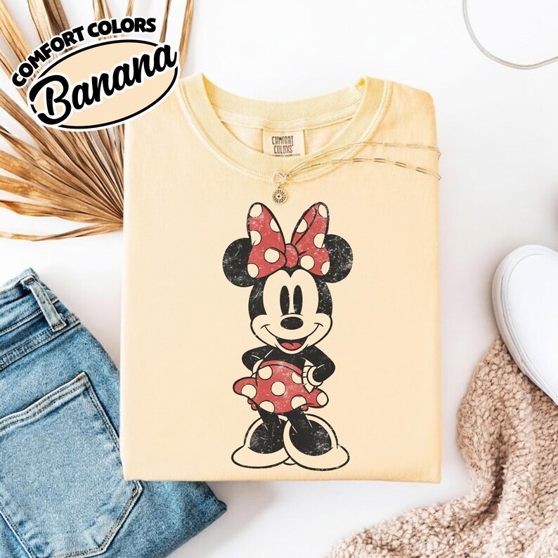 May include: A pale yellow t-shirt with a distressed Minnie Mouse graphic in a red polka dot dress and bow. The shirt has the words "Comfort Colors Banana" in a black oval. The shirt is shown with blue jeans and a white trainer.