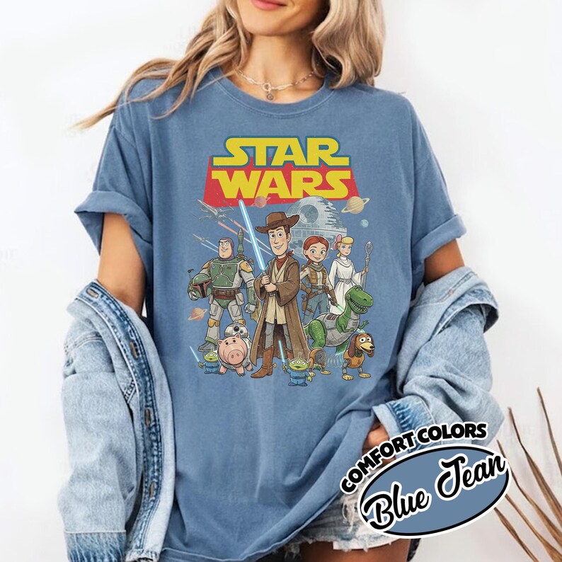 May include: Blue t-shirt featuring a graphic of Star Wars characters, including Woody, Buzz Lightyear, and others. The Star Wars logo is in red and yellow. The shirt is labeled "Comfort Colors Blue Jean."