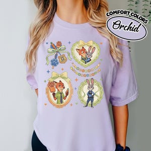 May include: A light purple t-shirt featuring Zootopia character graphics. The design includes Nick Wilde and Judy Hopps in ornate frames, a police badge, a paw-shaped popsicle, and the text Welcome to Zootopia, accented by small stars.