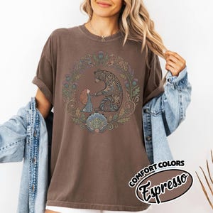 May include: Espresso-colored t-shirt featuring a circular design with a bear and a girl holding hands, surrounded by a floral wreath. The design is in shades of brown, green, and blue. The words "COMFORT COLORS Espresso" are printed on the bottom right.