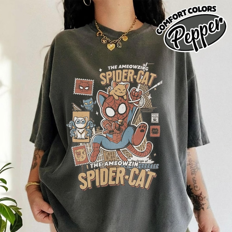 May include: Dark gray t-shirt featuring a cartoon Spider-Cat design. The graphic includes the text "THE AMEOWZING SPIDER-CAT" and a cat dressed as Spider-Man. The shirt also has the text "COMFORT COLORS" and "Pepper" in a circular design.
