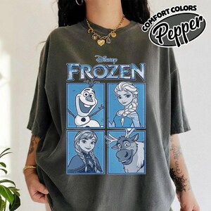 May include: Dark gray t-shirt featuring a Disney Frozen graphic. The graphic includes images of Olaf, Elsa, Anna, and Sven in blue squares. The text "FROZEN" is displayed above the characters. The shirt also has the text "COMFORT COLORS Pepper" in a circular design.