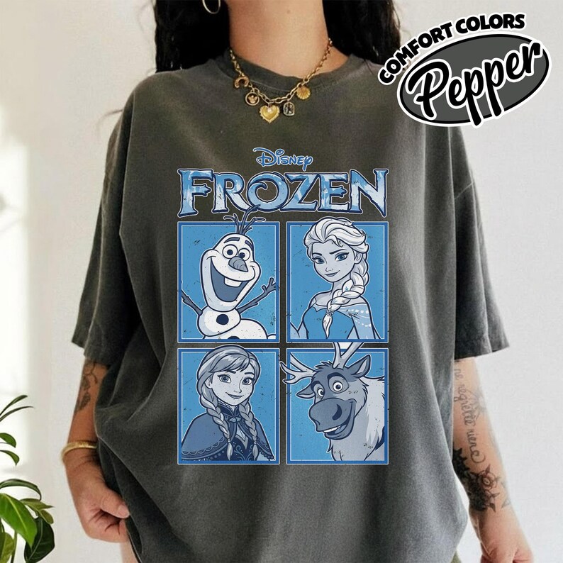 May include: Dark gray t-shirt featuring a Disney Frozen graphic. The graphic includes images of Olaf, Elsa, Anna, and Sven in blue squares. The text "FROZEN" is displayed above the characters. The shirt also has the text "COMFORT COLORS Pepper" in a circular design.