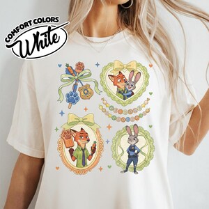 May include: A white t-shirt featuring a graphic print of Zootopia characters Nick Wilde and Judy Hopps. The design includes illustrations of the characters in ornate frames, a police badge, a carrot pen, and text that reads Welcome to Zootopia.