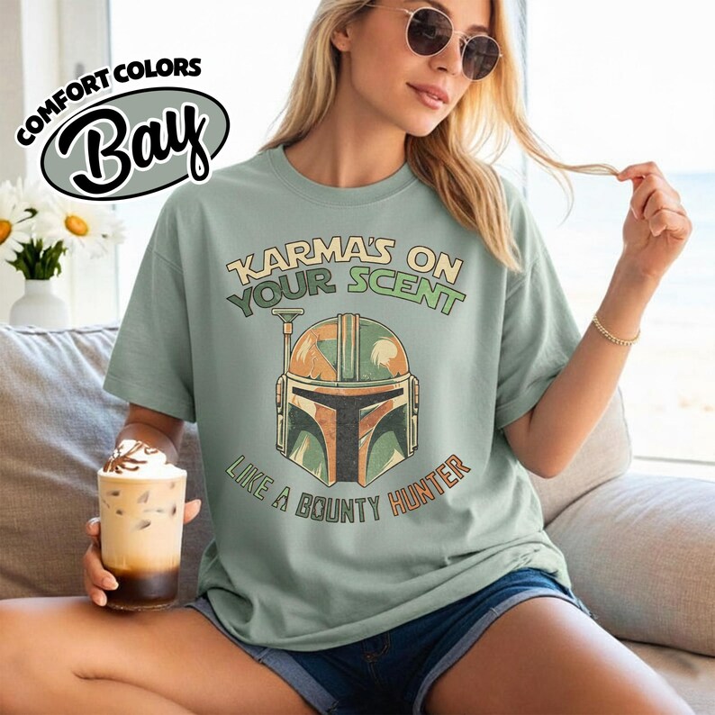 May include: A light green t-shirt with the text "Karma's on Your Scent Like a Bounty Hunter" and a graphic of a helmet. The shirt has a relaxed fit and is made of a soft material. The shirt is a Comfort Colors Bay brand.