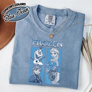 May include: A blue Comfort Colors t-shirt featuring characters from Disney's Frozen. The shirt displays Olaf, Elsa, Anna, and Sven in a four-panel design. A gold necklace rests on the shirt. The words "Comfort Colors" and "Blue Jean" are in the upper left corner.