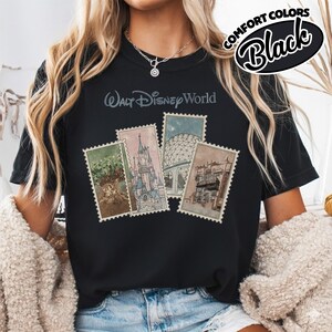 Op de afbeelding: Zwart T-shirt met de tekst "Walt Disney World" en vier postzegelontwerpen met Disney-attracties. Het shirt heeft ook de woorden "Comfort Colors" en "Black" in een cirkelvormig ontwerp.