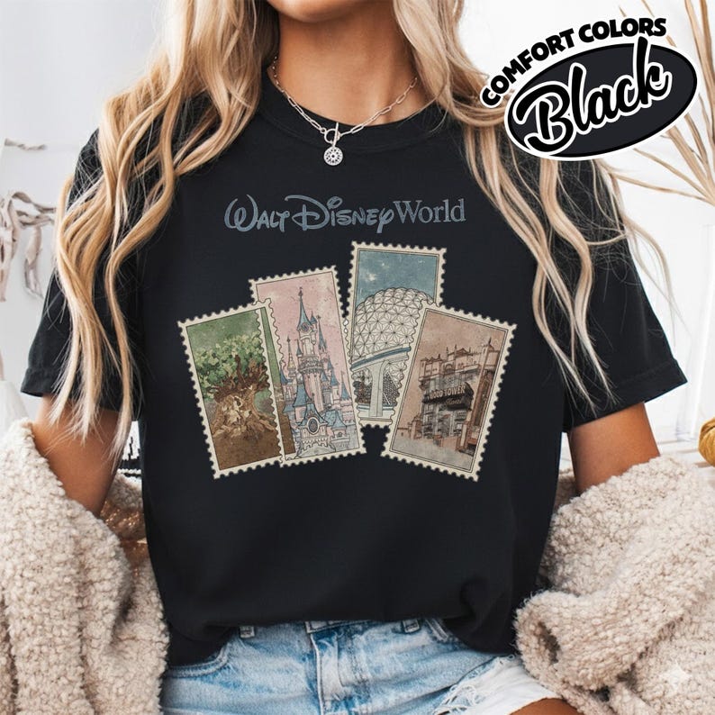 Op de afbeelding: Zwart T-shirt met de tekst "Walt Disney World" en vier postzegelontwerpen met Disney-attracties. Het shirt heeft ook de woorden "Comfort Colors" en "Black" in een cirkelvormig ontwerp.