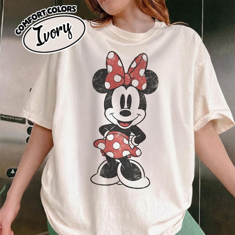May include: Ivory-coloured t-shirt with a distressed Minnie Mouse graphic. Minnie is in black, white, and red, with a polka-dot bow and dress. The shirt has short sleeves and a crew neck. The words "COMFORT COLORS" and "Ivory" are printed on the top left.