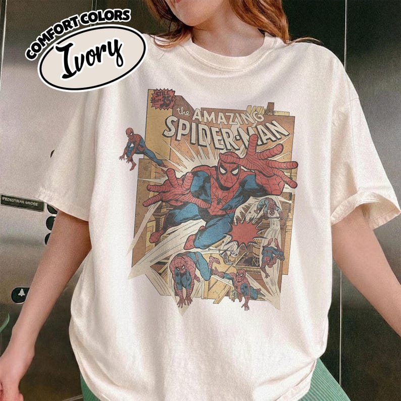 K&ouml;nnte beinhalten: Elfenbeinfarbenes T-Shirt mit Comic-Grafik von Spider-Man. Die Grafik enth&auml;lt den Text "The Amazing Spider-Man" und andere Comic-Elemente. Das T-Shirt hat kurze &Auml;rmel und einen Rundhalsausschnitt.