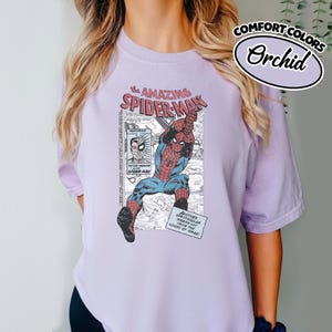 K&ouml;nnte beinhalten: Orchideenfarbenes T-Shirt mit einer Spider-Man-Comic-Grafik. Das Design enth&auml;lt den Text "The Amazing Spider-Man" und "Another Spectacular Slaan-Art From The House of Ideas!". Hergestellt aus Comfort Colors-Material.
