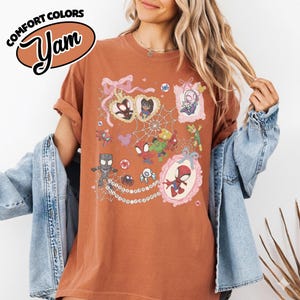 K&ouml;nnte beinhalten: Rostfarbenes T-Shirt mit einer farbenfrohen Grafik verschiedener Spider-Man-Charaktere in verzierten Rahmen und einem Spinnennetz-Design. Das Shirt hat die Worte "COMFORT COLORS" und "Yam" in einem braunen Oval.