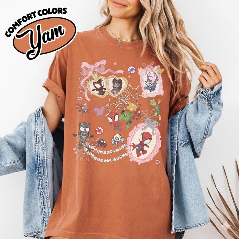 K&ouml;nnte beinhalten: Rostfarbenes T-Shirt mit einer farbenfrohen Grafik verschiedener Spider-Man-Charaktere in verzierten Rahmen und einem Spinnennetz-Design. Das Shirt hat die Worte "COMFORT COLORS" und "Yam" in einem braunen Oval.