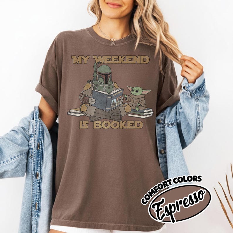 Może przedstawiać: Brązowa koszulka z napisem "MY WEEKEND IS BOOKED" i ilustracją postaci w zbroi czytającej książkę z małą zieloną istotą. Koszulka jest częścią kolekcji Comfort Colors.