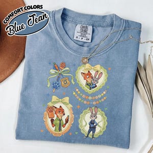 May include: A blue jean colored t-shirt featuring Zootopia character graphics. The design includes Nick Wilde and Judy Hopps in ornate frames, a police badge, a carrot, and a paw print. The shirt has a Comfort Colors tag at the neck.
