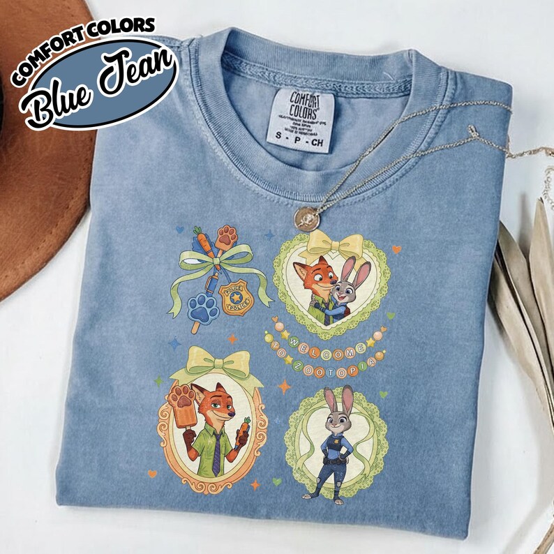May include: A blue jean colored t-shirt featuring Zootopia character graphics. The design includes Nick Wilde and Judy Hopps in ornate frames, a police badge, a carrot, and a paw print. The shirt has a Comfort Colors tag at the neck.