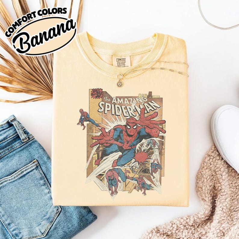 K&ouml;nnte beinhalten: Ein hellgelbes T-Shirt mit einer Vintage-Comic-Illustration von Spider-Man. Die Grafik zeigt Spider-Man in Aktion mit den Worten "The Amazing Spider-Man". Das Shirt tr&auml;gt die Aufschrift "Comfort Colors" und "Banana".