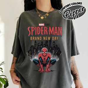 Puede incluir: Una camiseta gris carb&oacute;n con un gr&aacute;fico de Spider-Man agachado frente al horizonte de una ciudad. El texto dice Marvel Studios Spider-Man Brand New Day. La camiseta tiene un lavado estilo vintage, mangas cortas y cuello redondo.