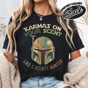 May include: Black t-shirt with the text "Karma's on Your Scent Like a Bounty Hunter" and a graphic of a Mandalorian helmet in shades of green, brown, and orange. The shirt also has the words "Comfort Colors" and "Black" in a circular design.