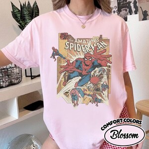 K&ouml;nnte beinhalten: Hellrosa T-Shirt mit Comic-Grafik von Spider-Man. Die Grafik enth&auml;lt den Text "The Amazing Spider-Man" und mehrere Illustrationen des Superhelden. Das Shirt ist mit "Comfort Colors Blossom" gekennzeichnet.