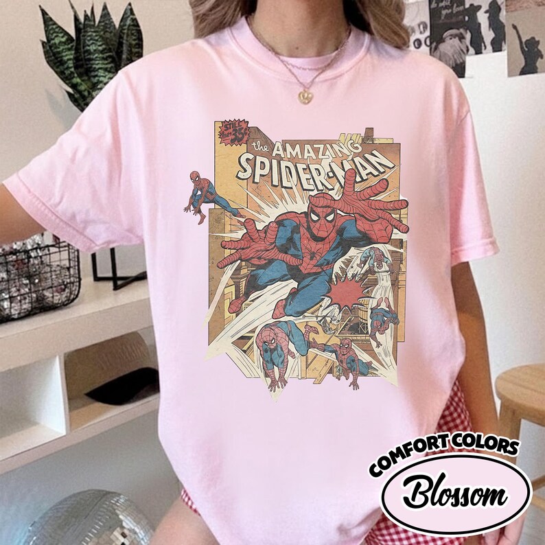 K&ouml;nnte beinhalten: Hellrosa T-Shirt mit Comic-Grafik von Spider-Man. Die Grafik enth&auml;lt den Text "The Amazing Spider-Man" und mehrere Illustrationen des Superhelden. Das Shirt ist mit "Comfort Colors Blossom" gekennzeichnet.
